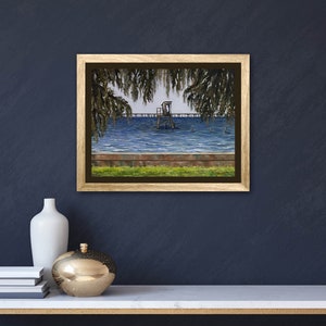 Pu&ograve; includere: Un dipinto incorniciato che rappresenta un molo di legno che si estende in un specchio d'acqua. Il molo ha una piccola struttura in legno. L'acqua &egrave; blu e ci sono alberi su entrambi i lati del molo. Il dipinto &egrave; in una cornice dorata.