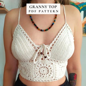 Può includere: Top all'uncinetto color crema con un design floreale e un laccio regolabile. Il top presenta un motivo quadrato nella parte inferiore e un bordo smerlato. Il testo "GRANNY TOP PDF PATTERN" è visualizzato in alto.