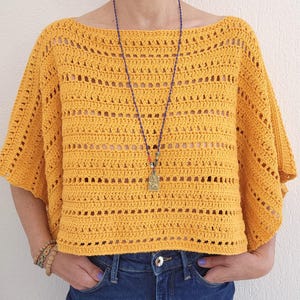 Puede incluir: Un top de ganchillo color amarillo mostaza con un amplio escote barco y mangas cortas y holgadas. La prenda está confeccionada con filas alternas de puntos macizos y patrones de malla calada, creando un aspecto texturizado y semitransparente.