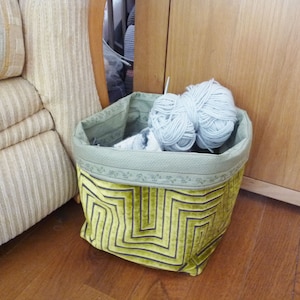 May include: A green and yellow fabric storage basket with a geometric pattern. The basket is filled with a ball of light blue yarn.