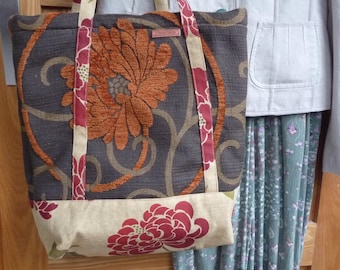 May include: A tote bag with a large orange floral design on a dark gray background. The bag has beige straps with red floral accents and a beige bottom panel with a red floral pattern. A small leather tag is attached to the strap.
