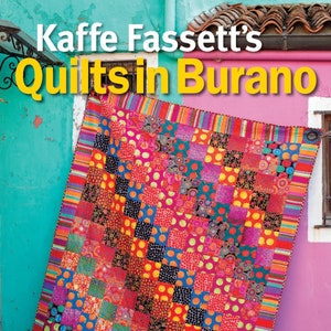 May include: A colorful quilt with a patchwork design featuring squares of different fabrics with polka dots, circles, and floral patterns. The quilt is hanging on a wall with a bright turquoise and pink background. The text "Kaffee Fassett's Quilts in Burano" is at the top of the image. The text "Designs inspired by a Venetian island" is at the bottom of the image.