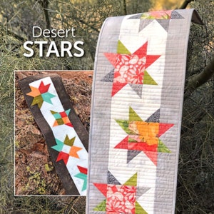 May include: A table runner with a gray background and a row of red, orange, and green stars. The stars are made of fabric with a floral pattern. The table runner is called "Desert Stars" and is by Atkinson Designs.