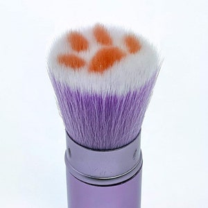 May include: A purple retractable makeup brush with a white and orange paw print design on the brush head.