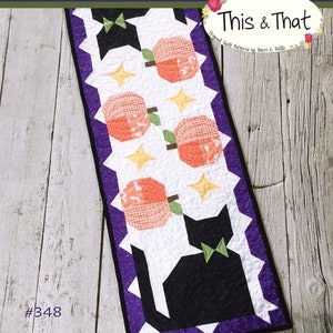 May include: A quilted table runner with a Halloween theme. The design features black cat silhouettes with green bow ties, orange pumpkins, and yellow stars on a white background. The runner is bordered with purple fabric and includes the text "Scaredy Cat" and "This & That".