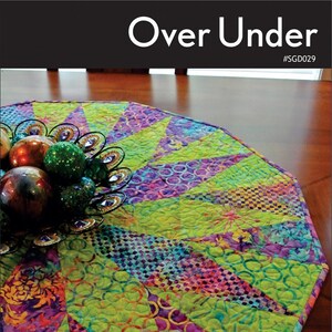 May include: A colorful quilted table runner with a green, purple, and pink floral pattern. The runner is made in a circular shape with a scalloped edge. The text "Over Under #SGD029" is printed on the top of the image. The text "Swirly Girls" is printed in a circle at the bottom of the image. The text "3 sizes for Over your table or Under your tree!" is printed below the logo.