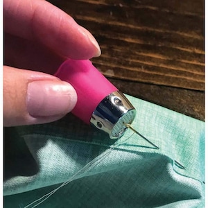 May include: A sewing thimble featuring a bright pink plastic top and a metallic silver base with a dimpled surface. The thimble is positioned over a piece of light green fabric, with a thin metal sewing needle and white thread visible.