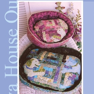 May include: Two quilted pet beds in a variety of colors and patterns. The smaller bed is pink and purple with a white cat design. The larger bed is green, blue, and purple with a white dog design. The text "Pieceful Pet Beds" is at the top of the image. The text "Java House Quilts" is at the bottom of the image.