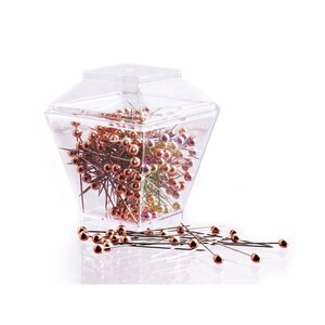 May include: A clear, faceted plastic container filled with numerous sewing pins featuring metallic copper-colored spherical heads. Several of these pins are scattered in front of the container, showcasing their long, thin, silver-toned metal shafts.