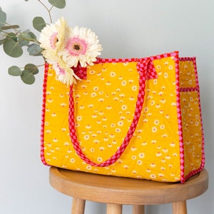 May include: A yellow quilted tote bag with red and pink checkered trim and daisy print. The bag has two handles and is sitting on a wooden stool.