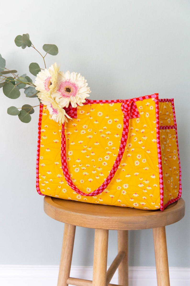 May include: A yellow quilted tote bag with red and pink checkered trim and daisy print. The bag has two handles and is sitting on a wooden stool.