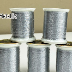May include: Five spools of metallic silver thread are stacked in a pyramid shape. The thread is wound on white plastic spools with gold labels. The word "Metallic" is printed in white on the top left.