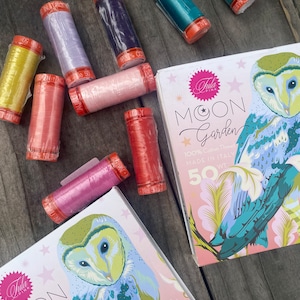 May include: A collection of Aurifil 50 weight cotton thread spools in yellow, lavender, dark purple, pink, coral, and teal, alongside two Tula Pink Moon Garden boxes featuring an owl illustration and the text Moon Garden 100% Cotton Thread.