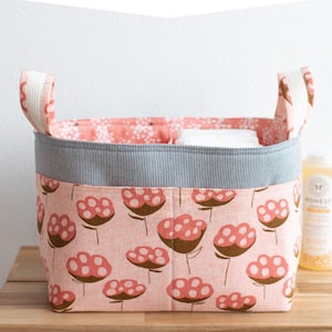 May include: A pink fabric basket with a divided lining and white handles. The basket has a floral print with brown stems and pink flowers with white dots. The text "DIVIDED BASKET" and "a structured basket with divided lining" is at the top of the image. The text "noodlehead" is at the bottom of the image.