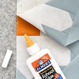 May include: A white bottle of Elmer's School Glue with a blue and orange label. The glue is washable and no-run. The bottle is sitting on a piece of fabric with a geometric pattern.