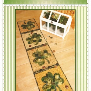 May include: A green and gold patchwork shamrock table runner with four shamrock designs. The table runner is on a wooden floor with a small white greenhouse in the background. The text "Patchwork Shamrock Table Runner" is at the bottom of the image.
