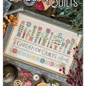 May include: A framed cross-stitch sampler titled "Garden of Quilts" with colorful floral designs. The sampler features a variety of flowers in pots and spools of thread. The text "Garden of Quilts" is stitched across the center. The design is by Lori Holt.