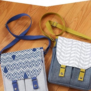 May include: Two gray messenger bags with different fabric patterns. The bag on the left has a blue and white geometric pattern. The bag on the right has a white leaf pattern. Both bags have yellow straps and silver buckles. The bags are on a wooden surface. #535 noodlehead