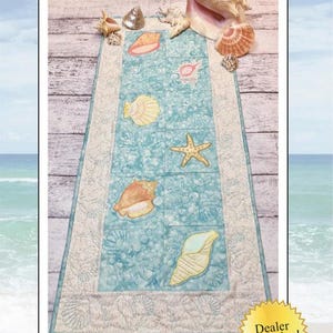 May include: A blue and white table runner with a seashell and starfish design. The text "On the Beach Table Runner" and "Custom quilting & machine appliqué" is at the top of the image. The text "AMÉLIE SCOTT DESIGNS" is at the bottom of the image. A yellow star with the text "Dealer Exclusive!" is in the lower right corner of the image.