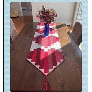 May include: A red and white quilted table runner with a tassel on the end. The runner is on a wooden table with a bouquet of pink and white flowers in a blue vase. The text "Love on the Run-ner" is at the top of the image and "Quilted Garden Designs by Janice Belling" is at the bottom.
