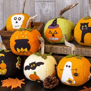 May include: A collection of handcrafted felt pumpkins with Halloween-themed designs. The pumpkins feature various faces, including a skull, jack-o'-lantern, cat, and ghost. Each pumpkin has a natural stem and is in shades of orange, green, black, and white.