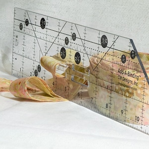 May include: A clear acrylic Add-A-Binding ruler by CM Designs, Inc. featuring black measurement markings and diagonal lines, positioned against a roll of light-colored fabric with a subtle pink and yellow mottled pattern for sewing projects.