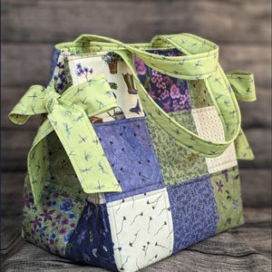May include: A green and purple patchwork tote bag with a green fabric handle and a bow. The bag has a floral pattern and dragonfly print.