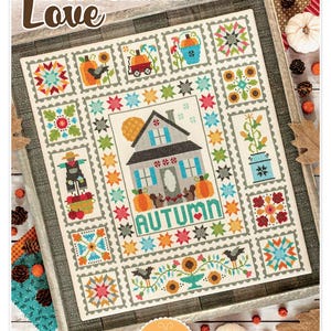 May include: A framed cross-stitch sampler with the words "Autumn Love" at the top. The sampler features a house, pumpkins, and other fall-themed motifs. The word "AUTUMN" is stitched in green. Designed by Lori Holl.