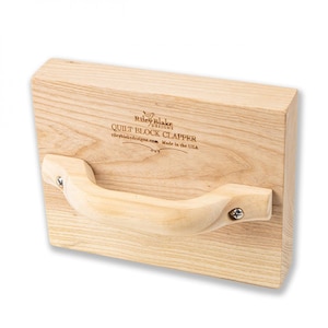 May include: A wooden quilt block clapper with a light wood handle attached with screws. The clapper is rectangular with the text "Riley Blake Designs QUILT BLOCK CLAPPER rileyblakedesigns.com Made in the USA" printed on the surface.