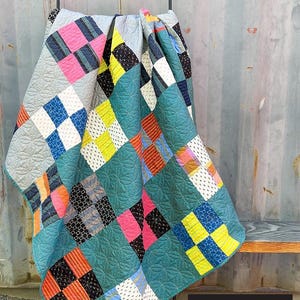 May include: A colorful patchwork quilt with a teal background and squares of pink, yellow, blue, and black. The quilt is draped over a black metal rack. The text "Norma's Patchwork" is at the top.