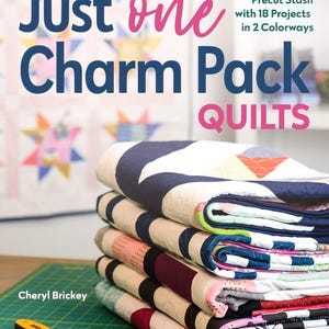 May include: A stack of colorful quilts with various patterns, including geometric and floral designs. The book cover reads "Just One Charm Pack Quilts" with the author's name, Cheryl Brickey, and a rotary cutter in the foreground.