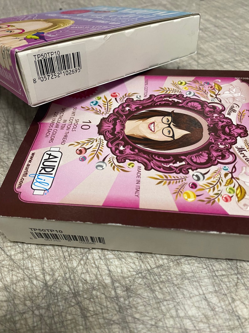 May include: A box of 70 spools of 50 weight cotton thread in ten spectacular colors. Each spool contains 220 yards of thread. The box is brown with a pink and white design featuring a woman with glasses. The text "Aurifil" is written in blue on the box. The box has a barcode with the product code TP50TP10.