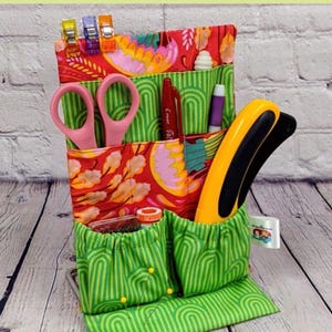 May include: A fabric organizer for sewing supplies, featuring multiple pockets and compartments. The organizer is approximately 5.5 x 8.5 inches. It has a green and red floral pattern, with pink scissors and other sewing tools visible. The text "SEW ORGANIZED" is displayed.