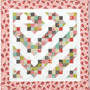 Pu&ograve; includere: Una trapunta patchwork con sfondo bianco e un motivo di quadrati colorati disposti a chevron. La trapunta &egrave; bordata con una stampa floreale rosa.