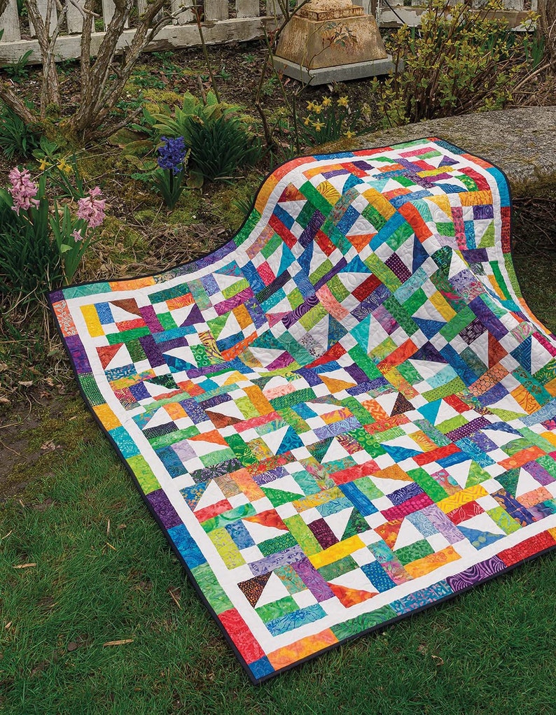 May include: A colourful patchwork quilt with a geometric pattern in shades of green, blue, orange, purple, and white. The quilt is made of various fabrics and is laid out on a grassy lawn.