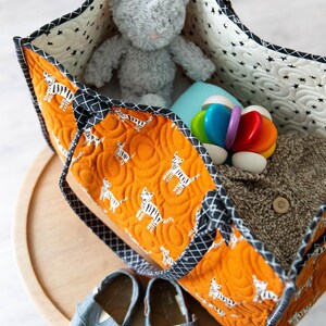 May include: A large orange and black tote bag with a zipper closure and two handles. The bag is filled with a gray stuffed animal, a blue blanket, a wooden toy, and a brown sweater.