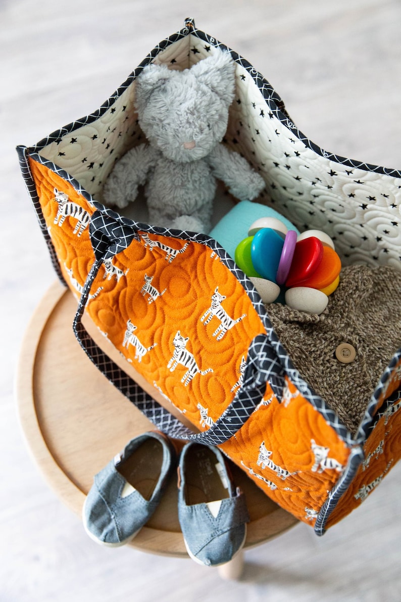 May include: A large orange and black tote bag with a zipper closure and two handles. The bag is filled with a gray stuffed animal, a blue blanket, a wooden toy, and a brown sweater.