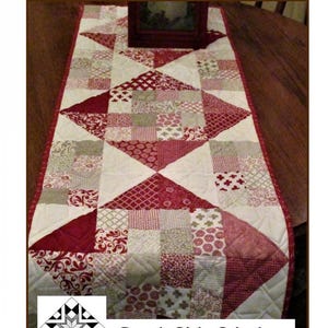May include: A quilted table runner with a geometric pattern in shades of red, white, and beige. The runner is 16 inches wide and 48 inches long. The text "Charming Road to Nowhere" and "Creek Side Stitches #288" is printed on the image.