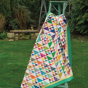 May include: A colourful quilt with a geometric pattern of triangles in various shades of blue, green, yellow, orange, pink, and purple. The quilt is draped over a wooden stool with a green finish. The quilt is displayed outdoors on a grassy lawn.