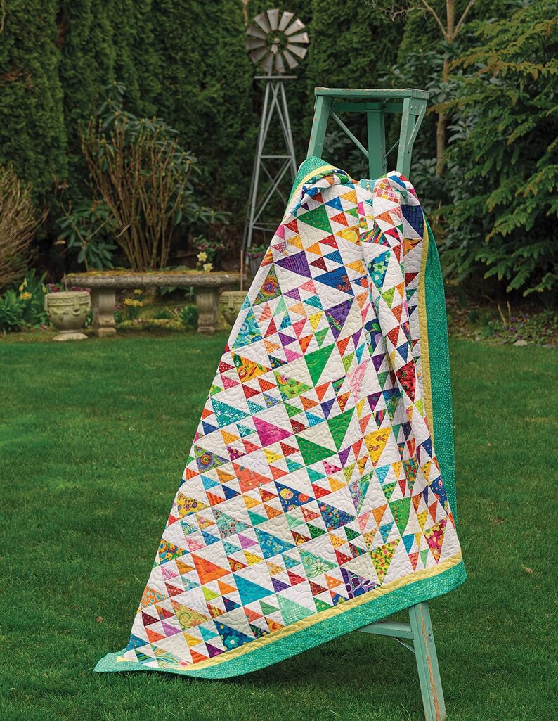 May include: A colourful quilt with a geometric pattern of triangles in various shades of blue, green, yellow, orange, pink, and purple. The quilt is draped over a wooden stool with a green finish. The quilt is displayed outdoors on a grassy lawn.