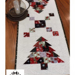 May include: A white quilted table runner with a Christmas tree design made of red, black, and green fabric squares. The runner is 16 1/2 inches by 40 inches and features a quilted design with a star in the corner. The text "Tis the Season" and "Creek Side Stitches #349" is printed on the runner.