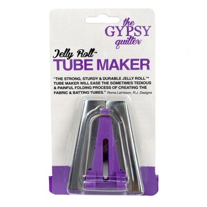 May include: A Jelly Roll Tube Maker, a quilting tool, is displayed on a white background. The tool is silver and purple, with the text "Jelly Roll Tube Maker" and "the GYPSY quilter" printed on the packaging. The packaging also includes a quote about the tool's function.