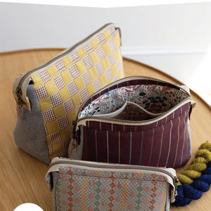 May include: Three zippered pouches in different sizes and patterns. The largest pouch is yellow and white checkered, the middle pouch is burgundy with white stripes, and the smallest pouch is light blue with a geometric pattern. The pouches are on a wooden surface with a pencil and yarn.
