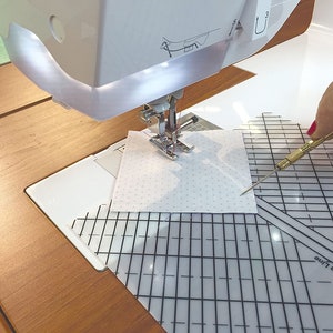 May include: A white fabric square with a small, white polka dot pattern is being sewn on a sewing machine. A clear ruler with 1/4 inch seam lines is visible underneath the fabric. A seam ripper is being held by a hand to the right of the fabric.