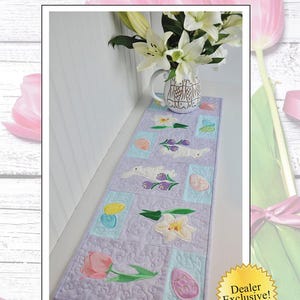 May include: A purple and white quilted table runner with an embroidered Easter theme. The runner features bunnies, eggs, flowers, and a pitcher of lilies. The text "Dealer Exclusive!" is on a yellow starburst.