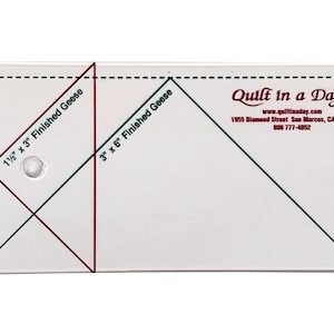 May include: A white plastic quilting ruler with black and green lines. The ruler has two sets of measurements for finished geese: 1 1/2 inches by 3 inches and 3 inches by 6 inches. The ruler also includes the text "Quilt in a Day" with the website address and phone number.