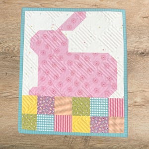 May include: A quilted wall hanging featuring a pink bunny silhouette with a floral pattern on a white background. The bottom border has a patchwork design with various colors and patterns, framed by a light blue border.