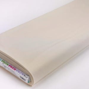May include: A roll of Kona cotton fabric in a light beige color. The fabric is neatly rolled and appears smooth. The end of the roll shows the Kona brand label with various colors.