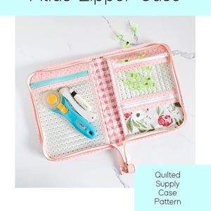 May include: Pink quilted Atlas Zipper Case with a floral print and multiple compartments. Includes a rotary cutter and other sewing tools. The case has a zipper closure and is labeled "Atlas Zipper Case" and "Quilted Supply Case Pattern".