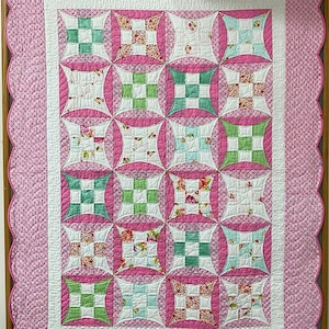 May include: A handmade quilt with a scalloped pink border and a central pattern of interlocking squares. The squares feature floral and geometric designs in pink, green, and white, creating a cozy and decorative textile.